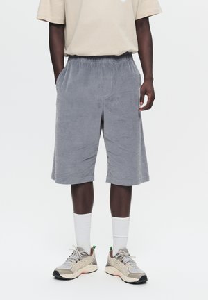 Man draagt beige t-shirt, grijze geribbelde knielange shorts, witte crew sokken en beige sportschoenen, staand op een witte achtergrond.