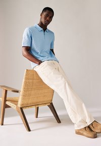 Světle modré polo tričko s texturovaným vzorem, krémové kalhoty a hnědé boty, sedící na tkané akcentové židli s dřevěnými nohami.