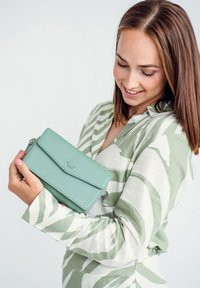 Mint green leather crossbody bag with a flap closure, featuring silver hardware and a logo, held by a person in a striped green shirt.