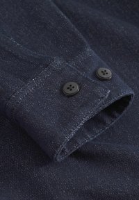 Puño de camisa de denim azul oscuro con dos botones negros, destacando una tela texturada con un brillo sutil y bordes cosidos.