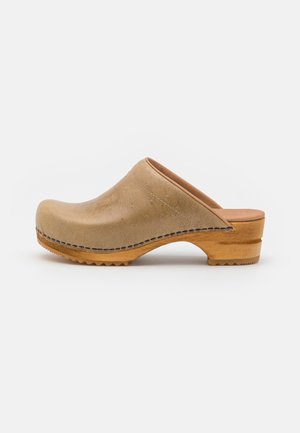 Sabot en cuir beige avec une texture lisse, une silhouette courbée et un talon en bois. Détail de couture notable le long de la couture supérieure.