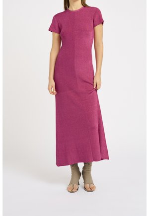 Vrouw draagt een aansluitende magentakleurige maxi-jurk met korte mouwen en beige open-toe hoge hakken tegen een effen achtergrond.