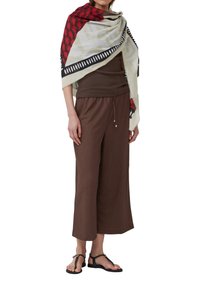 Châle léger crème avec des motifs géométriques rouges et noirs, superposé à un haut brun ajusté et un pantalon brun ample, associé à des sandales noires.