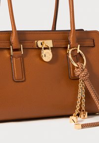 Bruine leren handtas met geprofileerd leer, goudkleurige hardware, een decoratieve sluiting, kettingaccenten en gestructureerde hengels. Bevat een logo-hanger en knoopdetail.