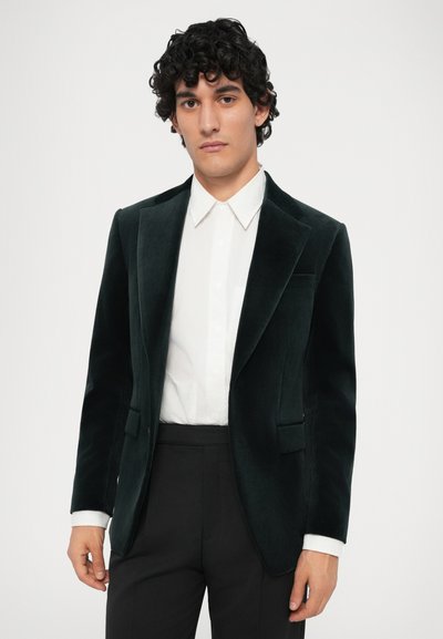 Tmavě zelen�ý sametový blazer s klopami, na jedno knoflíkové zapínání a dvěma předními kapsami, nošený přes bílé tričko a černé kalhoty.