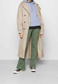 Gabardina beige, sudadera azul clara, blusa negra, pantalones acampanados verdes y zapatillas negras. Mezcla de tejidos con botones y detalles de cordón.