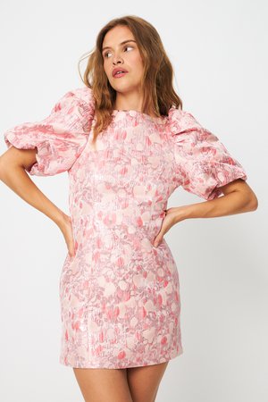 Γυναίκα με ροζ κοντό φόρεμα με σχέδια και φουσκωτά μανίκια στέκεται με τα χέρια στη μέση, κοιτώντας ελαφρώς προς το πλάι.