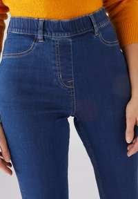 Jeans de mezclilla azul claro con una cintura alta, que cuentan con presillas para cinturón, bolsillos laterales y costuras naranjas en contraste. Textura suave, diseño ajustado.