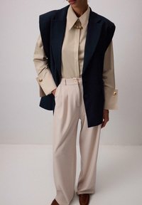 Tmavomodrý prešívaný blejzer s zlatými gombíkmi, béžová košeľa s golierom a krémové široké nohavice. Hladká látka s jemným leskom.