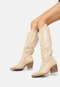 Botas de cuero beige hasta la rodilla con puntera cuadrada, cremallera lateral y un tacón de madera apilada. Textura suave con detalles mínimos.