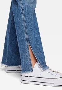 Jeans de mezclilla con dobladillos deshilachados y aberturas laterales, combinados con zapatillas de plataforma blancas con rayas negras en la suela.