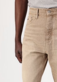 Vaqueros de mezclilla beige con cintura alta, bolsillos delanteros y un pequeño logo cerca del bolsillo. Textura suave con un corte clásico.