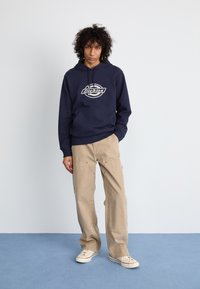 Hoodie marine avec un logo central, associé à un pantalon beige ample. Le hoodie possède une poche avant et des poignets côtelés. Des baskets complètent le look.