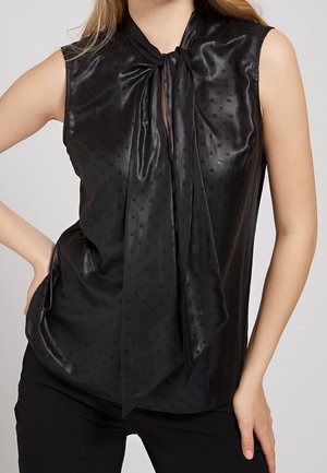 Ärmellose schwarze Satinbluse mit dezenten Polka-Dots und großer Schleife am Ausschnitt, getragen zu schwarzen Hosen.