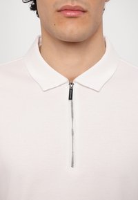 Cămașă polo albă cu detaliu de guler cu fermoar. Material neted, ușor, cu o textură subtilă și un zip argintiu.