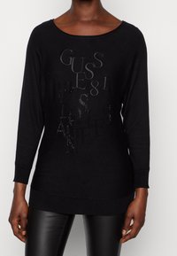 Suéter negro de manga larga con un escote amplio. Presenta un texto en relieve y brillante, "GUESS 81" y "LOS ÁNGELES." Material de punto suave.