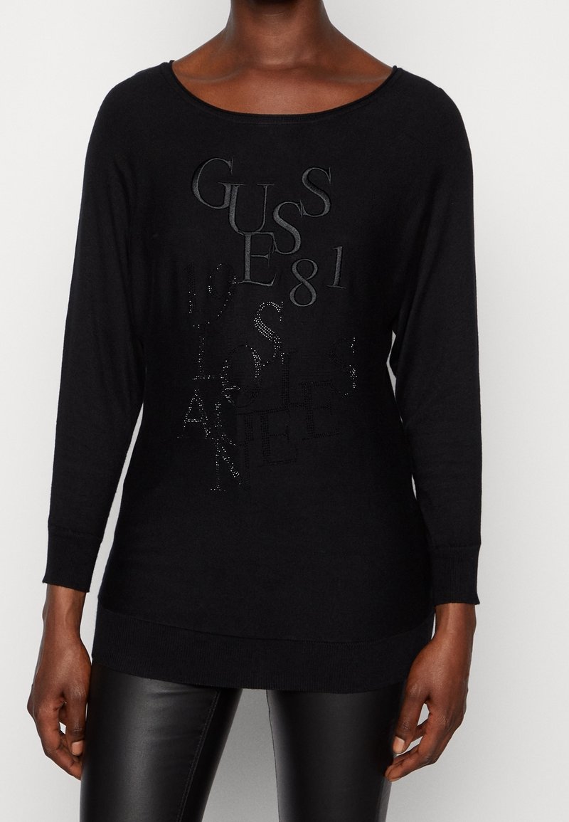 Suéter negro de manga larga con un escote amplio. Presenta un texto en relieve y brillante, "GUESS 81" y "LOS ÁNGELES." Material de punto suave.