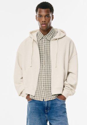 Beige zip-up hoodie with large front pockets over a green plaid shirt, featuring a collared neck. Worn with blue jeans. Smooth texture.