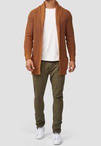 Brązowy sweter z dzianiny z kołnierzem szalowym, otwarty z przodu i z dwiema kieszeniami z przodu. Noszony na białej koszuli, w zestawieniu z oliwkowymi spodniami.