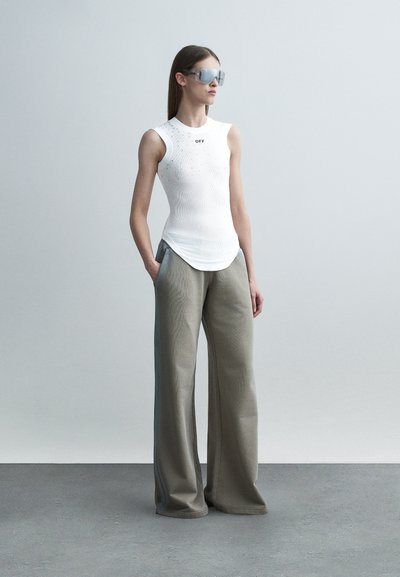 Γυναίκα που φοράει λευκό τοπ χωρίς μανίκια με το κείμενο «OFF», φαρδιά γκρι παντελόνια και μεγάλα αντανακλαστικά γυαλιά ηλίου, στέκεται με τα χέρια στις τσέπες.