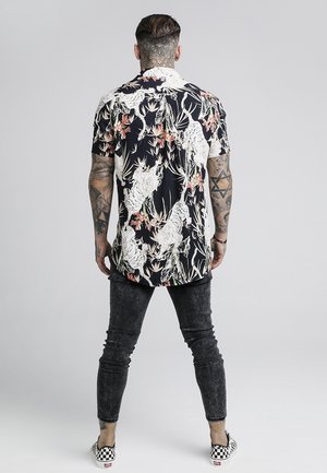 Mand med tatoverede arme og nakke iført en sort blomstret kortærmet skjorte, falmede sorte skinny jeans og ternede slip-on sko, stående med ryggen til.