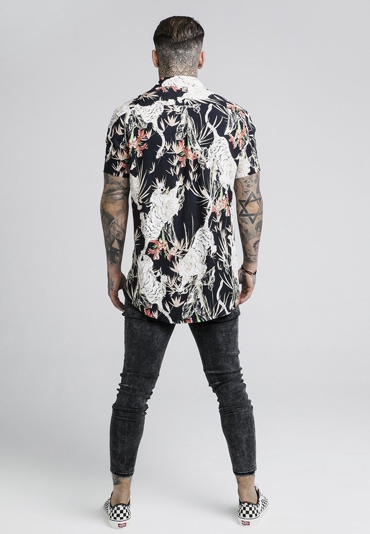 Homme aux bras et au cou tatoués portant une chemise noire à fleurs à manches courtes, un jean skinny noir délavé et des chaussures slip-on à carreaux, debout, vu de dos.