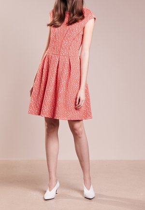 Woman wearing a knee-length, coral pink patterned dress with short sleeves and white low-heeled shoes, standing on a beige floor.