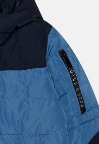 Pamušalotas švarkas su tamsiai mėlyna viršutine dalimi ir šviesiai mėlyna apatine dalimi, turi užtrauktuką ir etiketę "OTCH & SODA" ant rankovės.