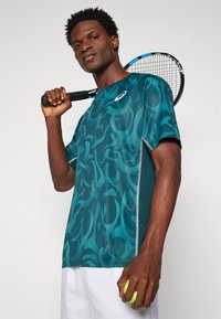 Teal sportlik särk, millel on keerduv muster, lühikesed varrukad ja kontrastsed külgpaneelid. Ühes käes hoiab ta tennisereketit ja teises tennisepalli.
