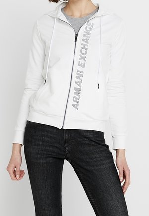 Femme portant un sweat à capuche blanc zippé avec le texte "ARMANI EXCHANGE" en diagonale sur le devant, associé à un jean noir et un T-shirt gris en dessous.