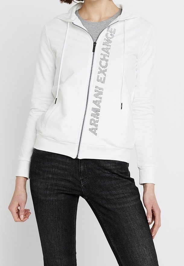Femme portant un sweat à capuche blanc zippé avec le texte "ARMANI EXCHANGE" en diagonale sur le devant, associé à un jean noir et un T-shirt gris en dessous.