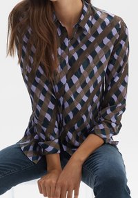 Femme assise, portant une chemise boutonnée à manches longues avec un motif à carreaux marron, violet et noir, et un jean bleu.
