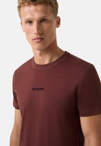 Vínová krátká trička s rukávem vyrobená z měkké bavlny, s kulatým výstřihem a černým natištěným logem "BOGNER" na prsou. Hladká textura.