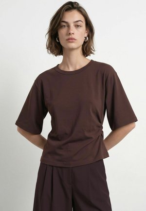 Frau mit kurzen braunen Haaren trägt ein dunkelbraunes oversized T-Shirt mit kurzen Ärmeln und passende lockere Hose vor einfarbigem Hintergrund.