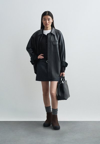 Chemise-veste oversized en cuir noir, jupe en denim foncé, bottes marron à talons avec semelles noires, chaussettes côtelées grises et sac à main noir.