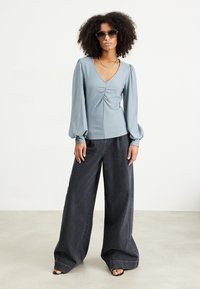 Top azzurro chiaro a maniche lunghe con scollo a V profondo e dettaglio arricciato, abbinato a pantaloni in denim nero a gamba larga, indossato con sandali neri.