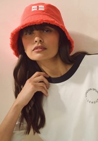 Czerwona chusta na głowę z postrzępionym rondem i logo Anglii. Model ma na sobie biały top z czarnymi akcentami i nadrukiem logo. Gładka faktura, casualowy styl.