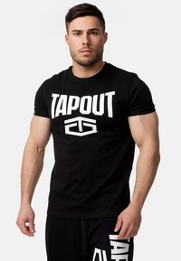 Melns kokvilnas T-krekls ar lielu baltu „TAPOUT” logo priekšpusē. Redzami īsie piedurknes un klasiskā apaļā kakla izgriezums.