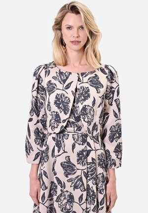 Femme blonde portant une robe beige et une veste courte assortie avec un grand motif floral noir, debout devant un fond uni clair.
