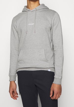Mann trägt einen hellgrauen Hoodie mit dem Schriftzug "LES DEUX" auf der Brust und schwarze Hose, steht vor einem schlichten hellen Hintergrund.