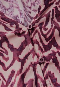 Blouse met knopen in een roze en paarse abstracte patroon, gemaakt van zachte stof met een V-hals en twee bijpassende knopen bij de kraag.