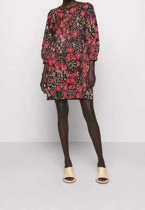 Kobieta ubrana w czarną sukienkę do kolan z czerwonymi kwiatowymi i leopardowymi wzorami oraz beżowe odkryte sandały typu klapki, stojąca na jednolitym tle.