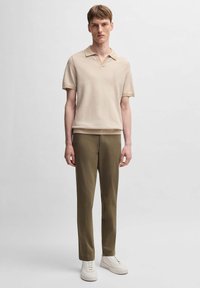 Polo de culoare bej, cu mânecă scurtă și textură cu caneluri, asortat cu pantaloni în culoare verde măsliniu și adidași albi. Design simplu, modern, cu o croială relaxată.