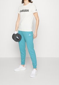 White t-shirt with black "adidas" logo, turquoise sweatpants with ankle cuffs, white sneakers, and a black weight plate held in one hand.