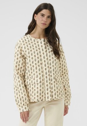 Veste crème matelassée avec motif floral, présentant un col rond, des manches longues et deux poches avant. Texture de tissu lisse.