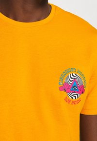 Žluté bavlněné tričko s kulatým výstřihem. Na přední straně se nachází barevná grafika: oko v trojúhelníku a text "Zdeformovaná realita Los York."