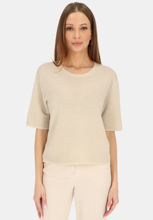 Kobieta w beżowej prążkowanej bluzce z krótkim rękawem i pasujących beżowych spodniach, stojąca na tle jednolitego jasnego tła.