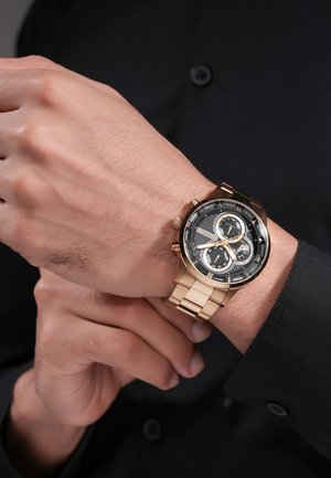 Mies, joka säätää mustan kellotaulun ja kolmen apukellon omaavaa kultaisen kronografikellon ranneketta mustan pitkähihahaisen paidan päällä.