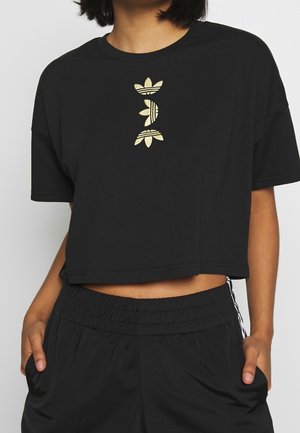 Osoba ubrana w czarny krótki T-shirt z pionowym złotym logo Adidas oraz czarne spodnie z elastycznym pasem, z rękami w kieszeniach.