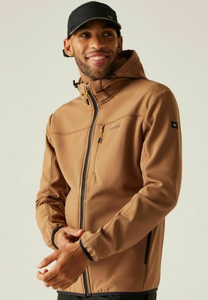 Mann, der lächelt, trägt eine braune Kapuzenjacke mit Frontreißverschluss und eine schwarze Mütze, vor einem schlichten hellen Hintergrund.